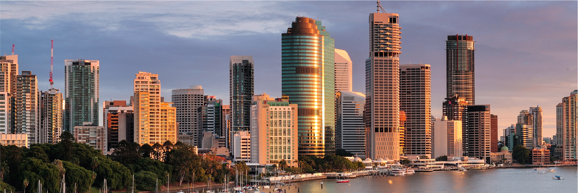 City skyline with tall buildings reflecting sunlight, waterfront with boats docked, and green trees in the foreground.